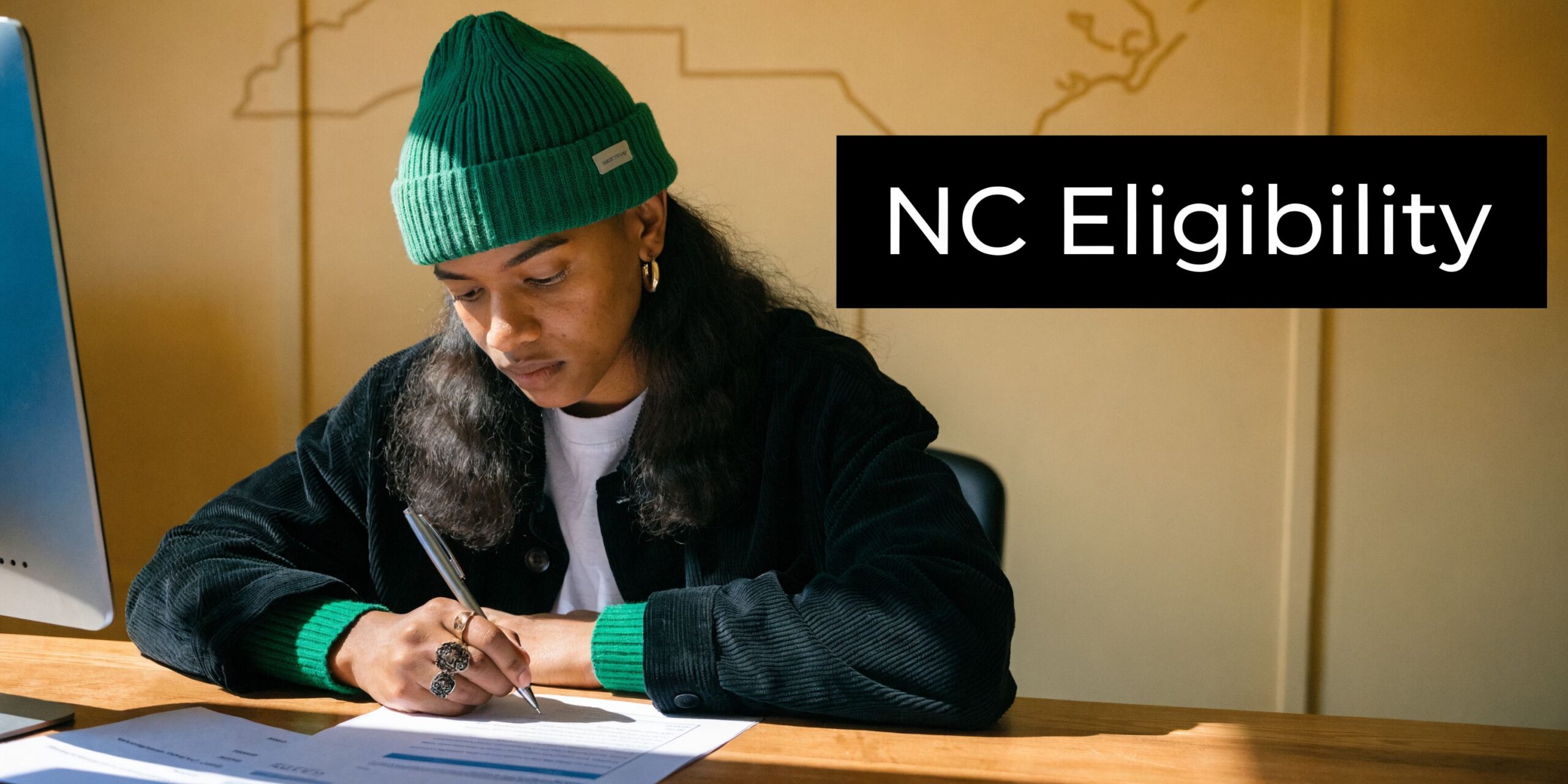 A person wearing a green beanie writing on documents at a desk under an NC Eligibility sign.
